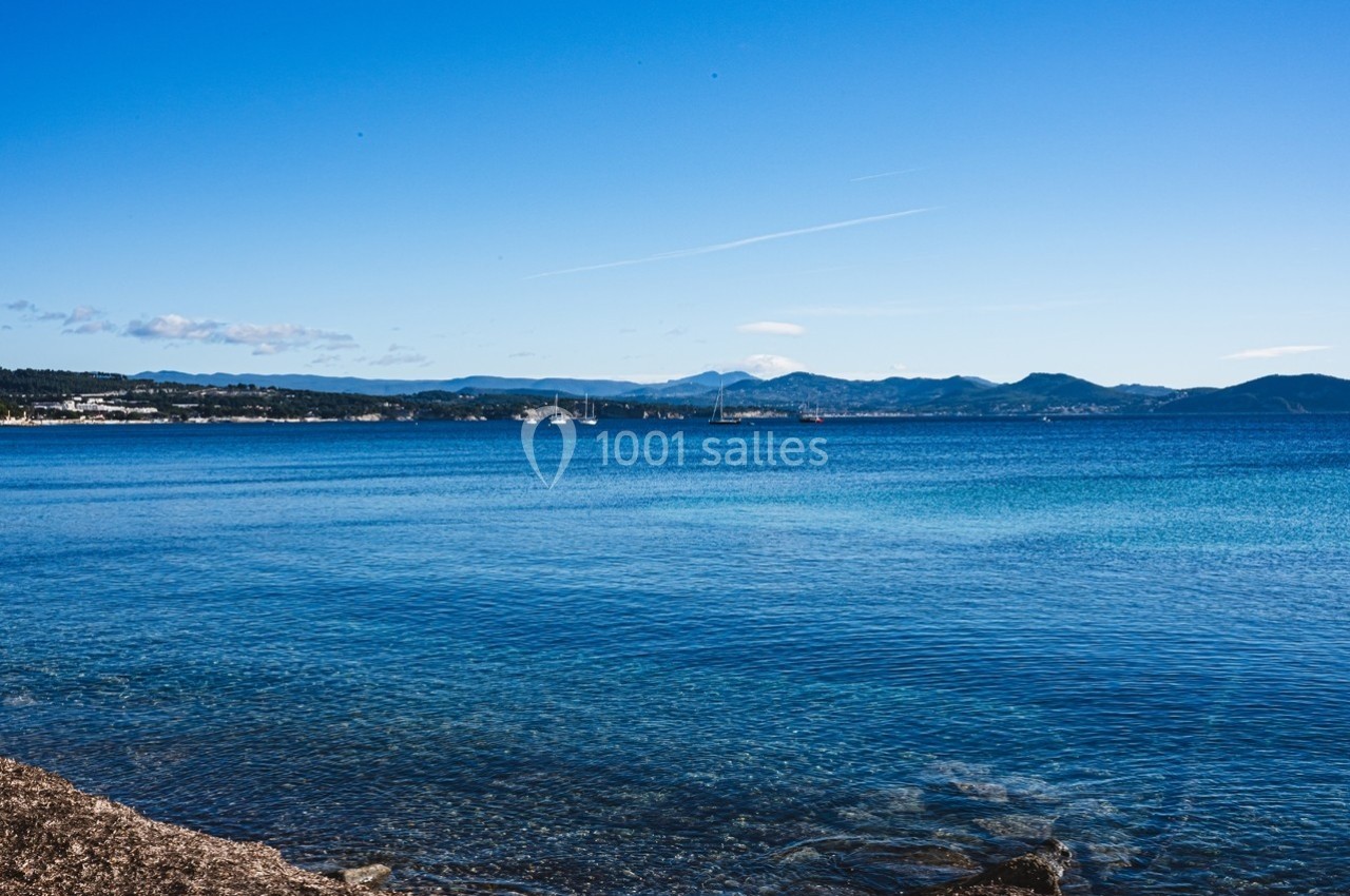 Vue sur une mer calme avec un rivage rocheux au premier plan et des montagnes à l'horizon sous un ciel dégagé.