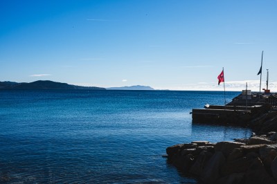 Vue d'un littoral avec une mer calme, un quai, des drapeaux et des collines à l'horizon sous un ciel dégagé.