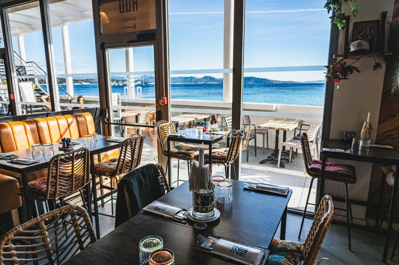 Salle de restaurant lumineuse avec vue sur la mer, tables en bois, chaises en rotin et grande baie vitrée.