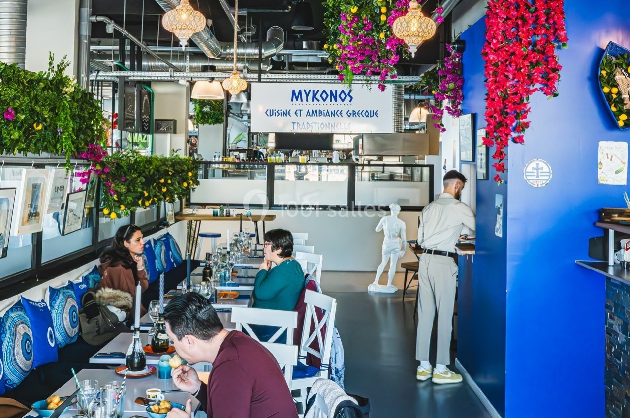 Salle de restaurant décorée de plantes suspendues, avec des clients attablés et un serveur près du comptoir.