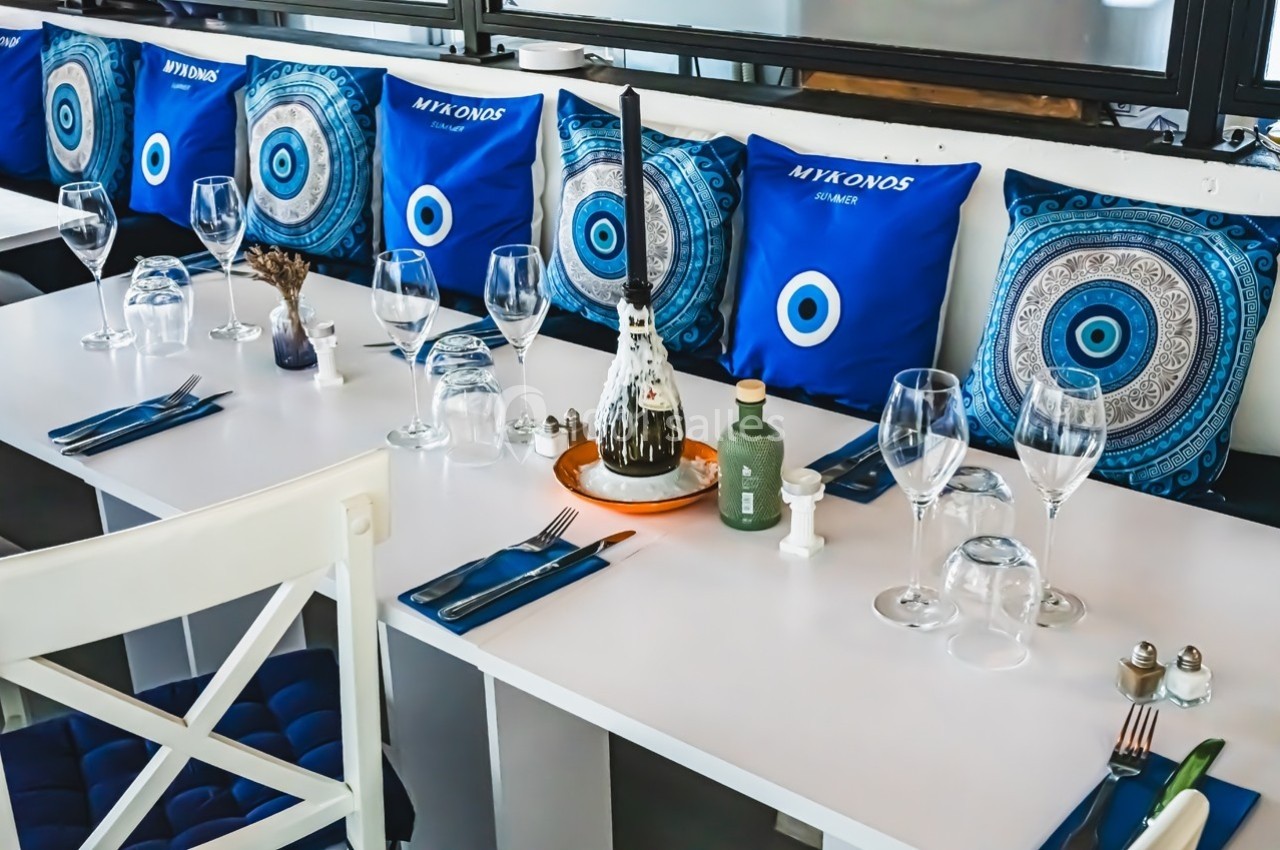 Table dressée avec des verres, couverts et bouteilles, entourée de coussins bleus décorés de motifs grecs.