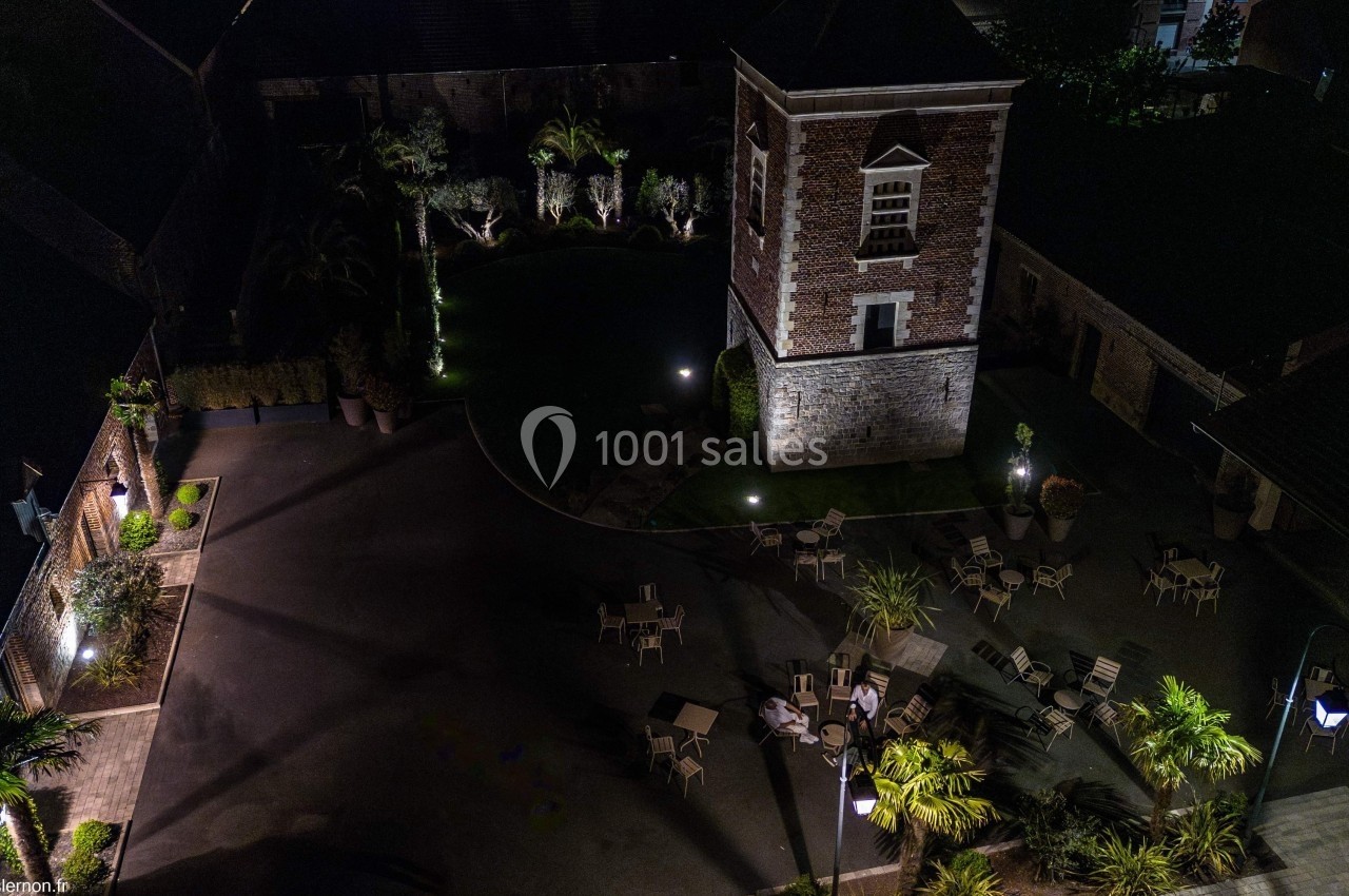 Vue aérienne nocturne d'une cour avec une tour en briques, des tables, des chaises et un éclairage paysager.