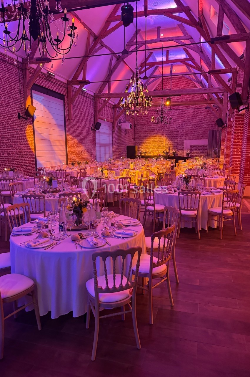 Salle de réception décorée avec des tables rondes dressées, éclairage chaleureux et poutres apparentes en bois.