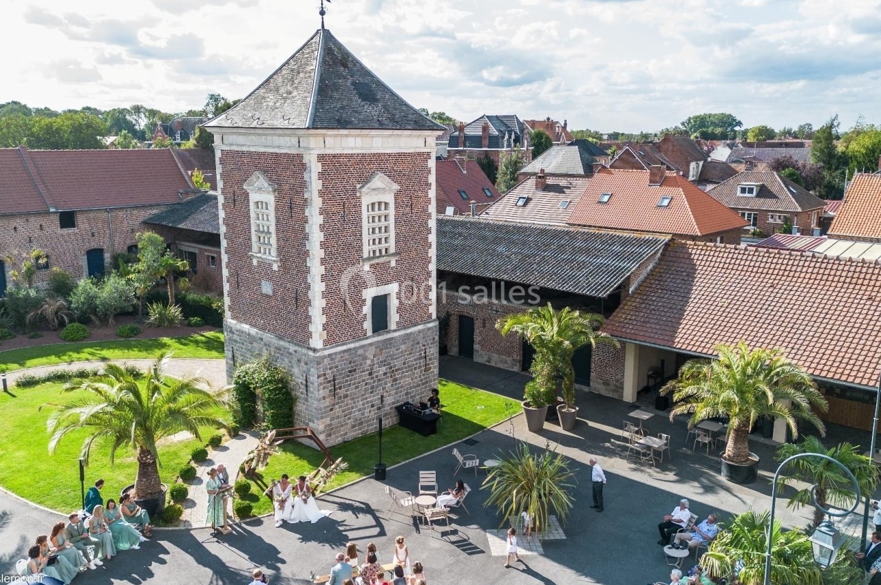Vue aérienne d'une cour avec une tour en briques, des bâtiments anciens, des palmiers et des personnes réunies.