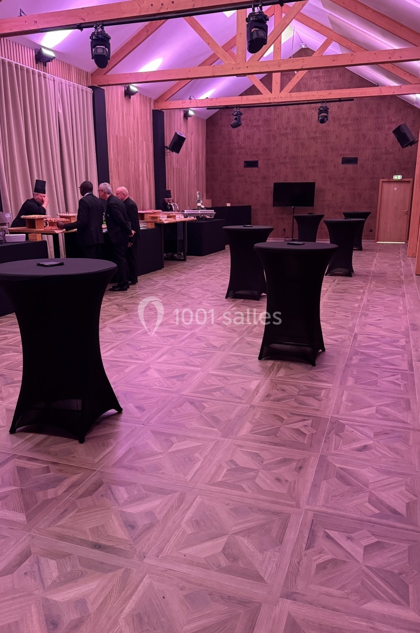 Salle de réception avec tables hautes noires, parquet clair et éclairage tamisé, quelques personnes au fond.