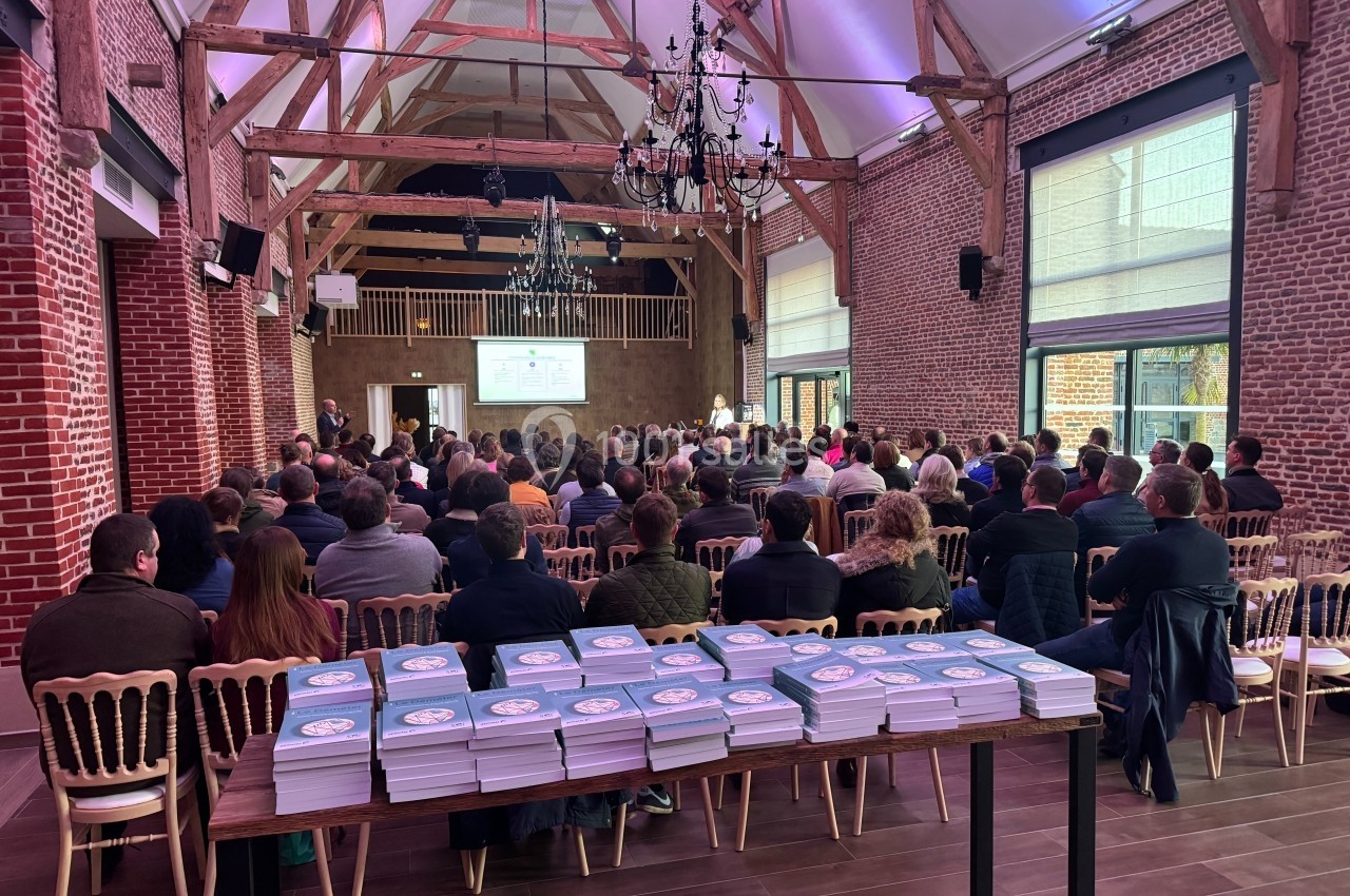 Salle de conférence en briques avec un public attentif, des présentations projetées et des brochures empilées au premier…