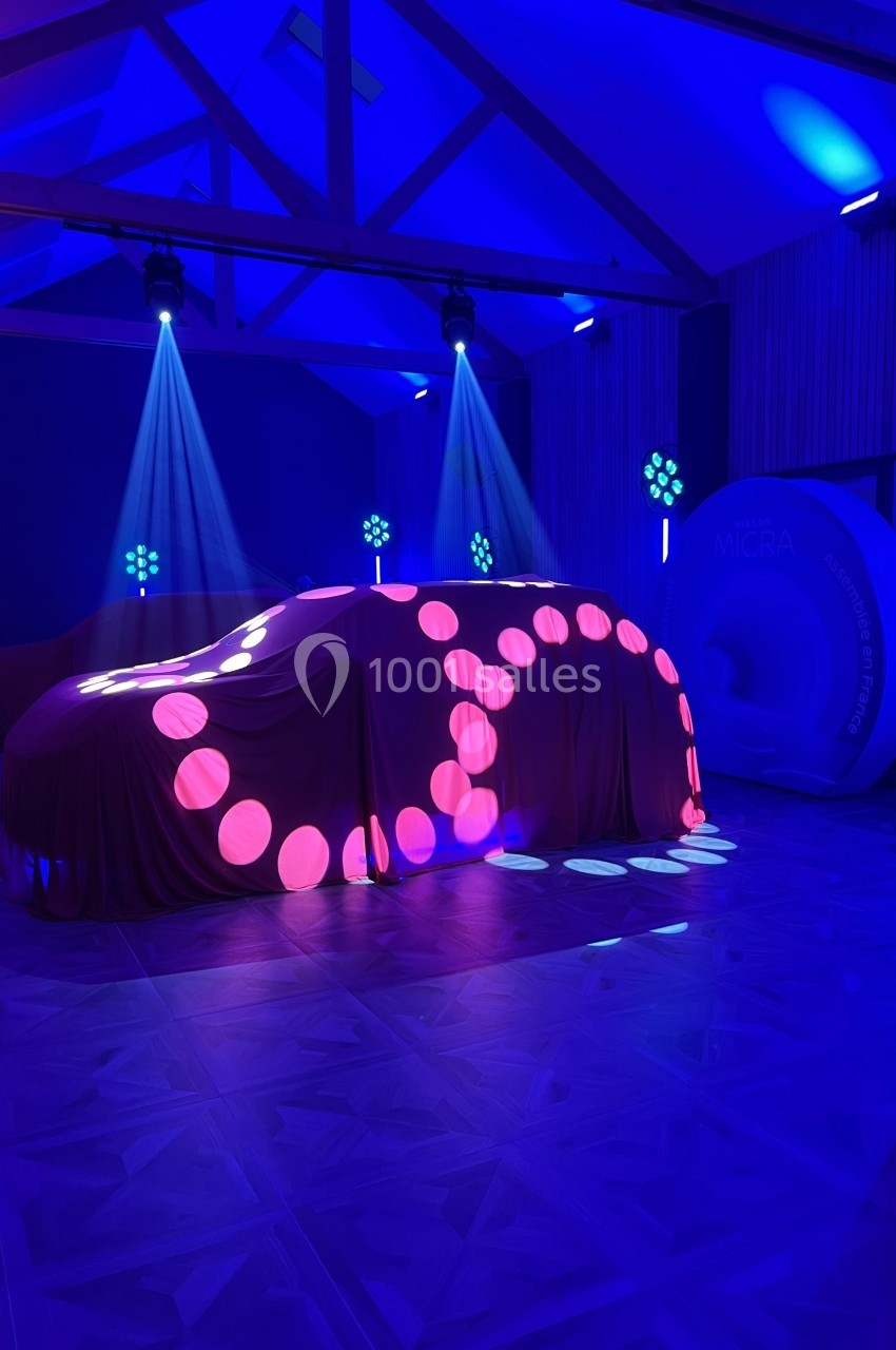 Voiture recouverte d'un drap illuminé par des motifs lumineux roses et bleus dans une salle éclairée en bleu.