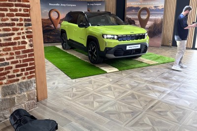 SUV vert exposé dans une salle avec parquet et murs en bois, accompagné de panneaux promotionnels en arrière-plan.