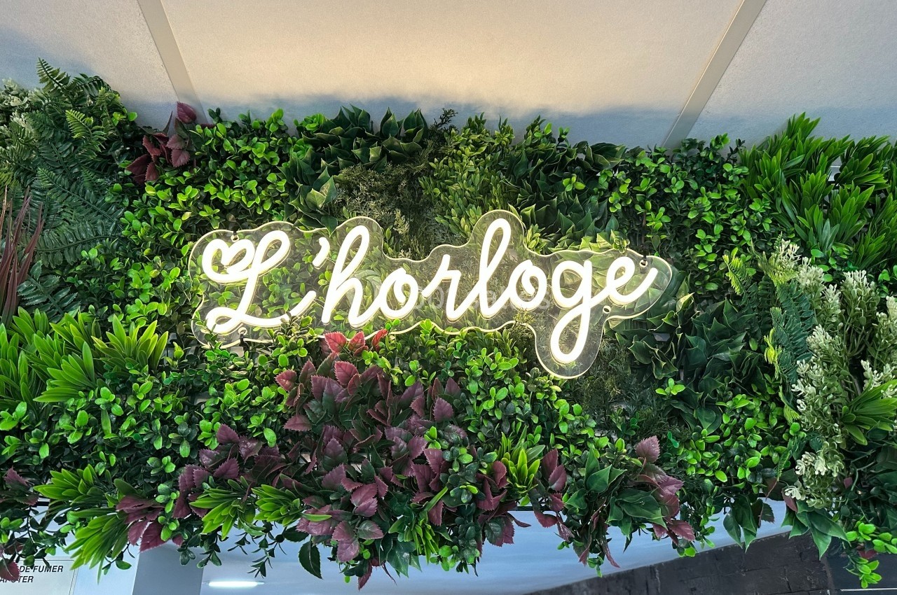 Enseigne lumineuse ’L'horloge’ entourée de feuillage vert et de fleurs décoratives.