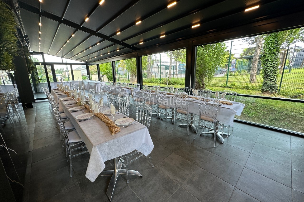 Salle de réception lumineuse avec grandes baies vitrées, tables dressées et vue sur un espace extérieur verdoyant.