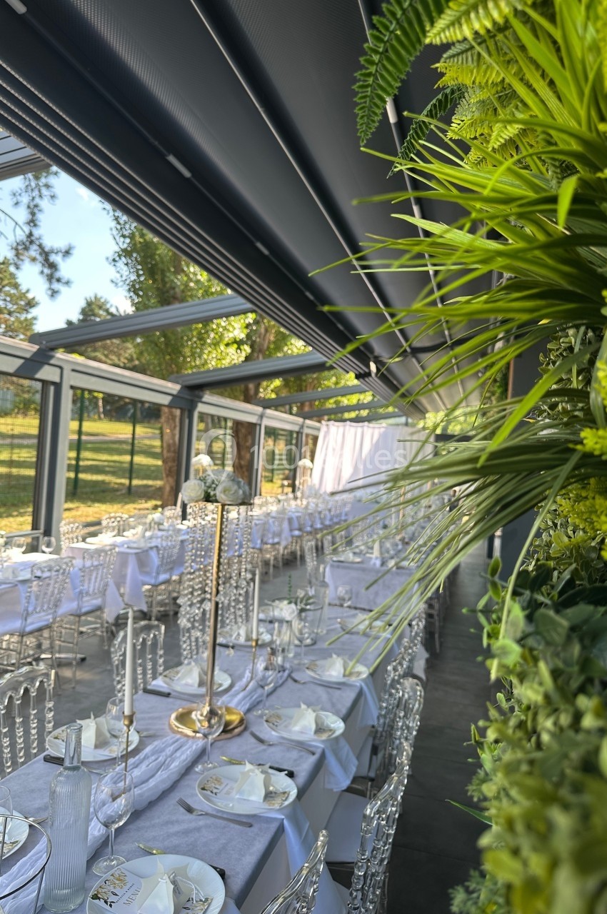 Salle de réception lumineuse avec tables décorées de nappes blanches, chaises transparentes et plantes vertes en arrière…