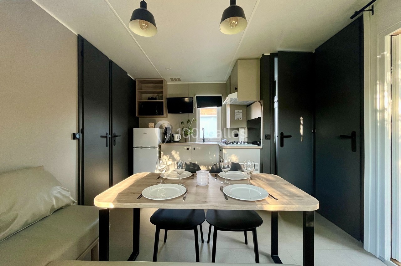 Salle à manger moderne avec table dressée pour quatre, cuisine équipée en arrière-plan et éclairage suspendu.