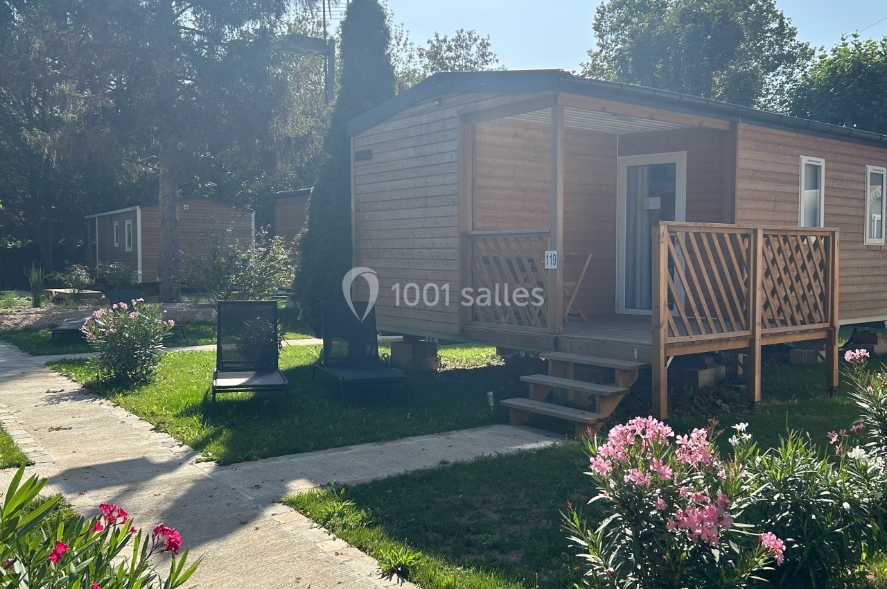 Mobil-home en bois avec terrasse, entouré de végétation, chaises longues et fleurs dans un cadre ensoleillé.