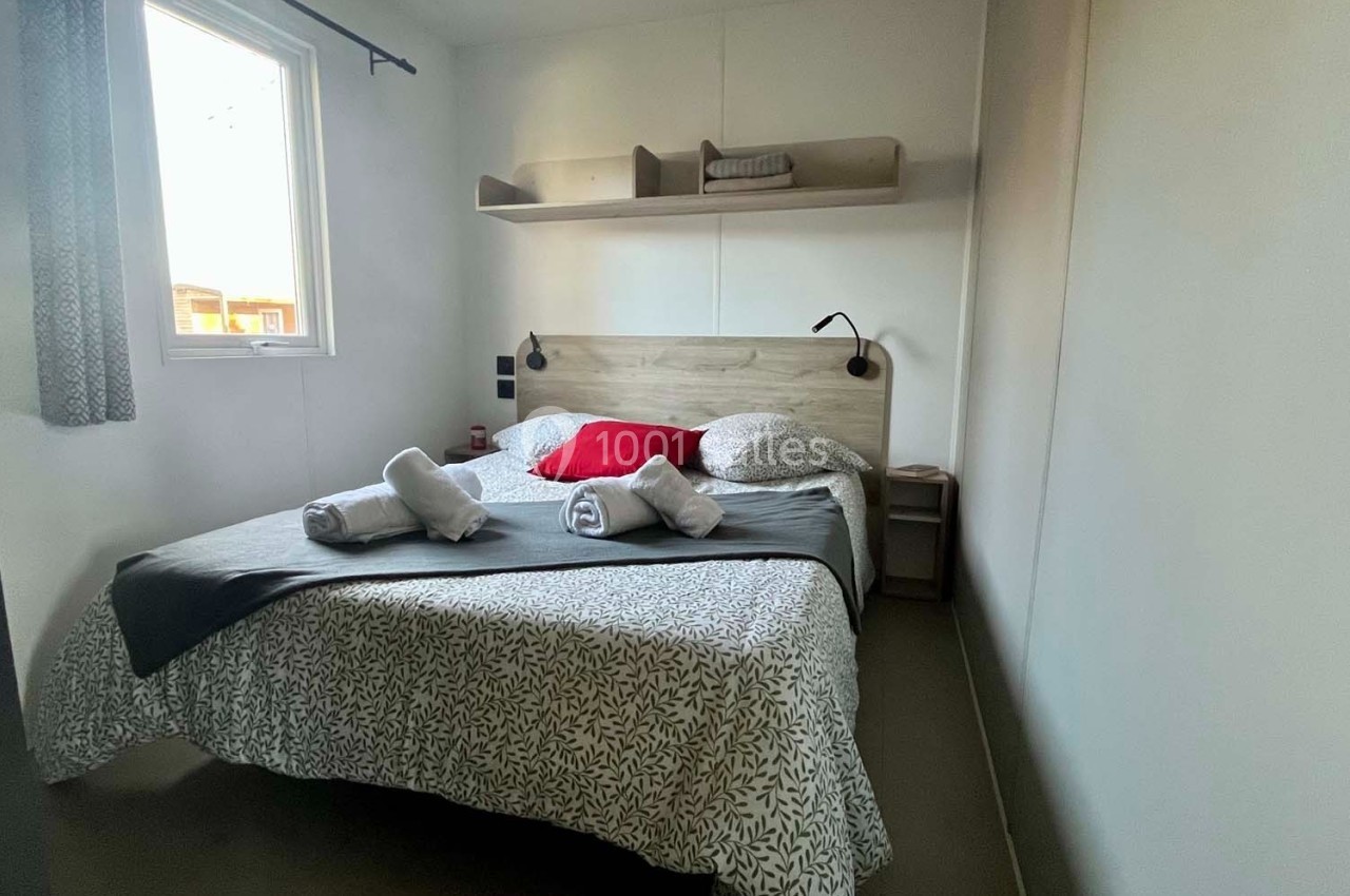 Chambre avec lit double, draps pliés, coussins rouges, étagère murale et fenêtre laissant entrer la lumière naturelle.