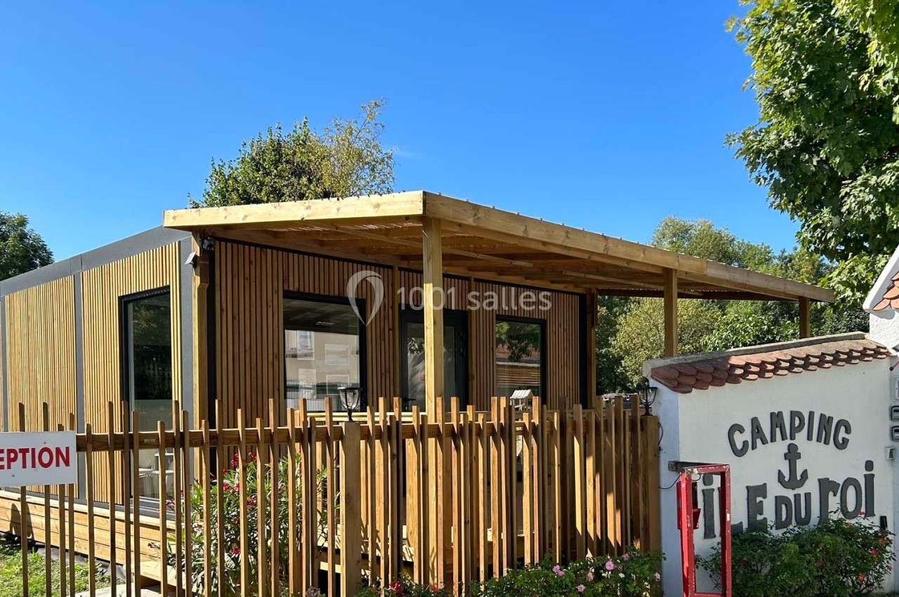 Bâtiment en bois moderne avec terrasse couverte, situé à l'entrée d'un camping entouré de verdure.