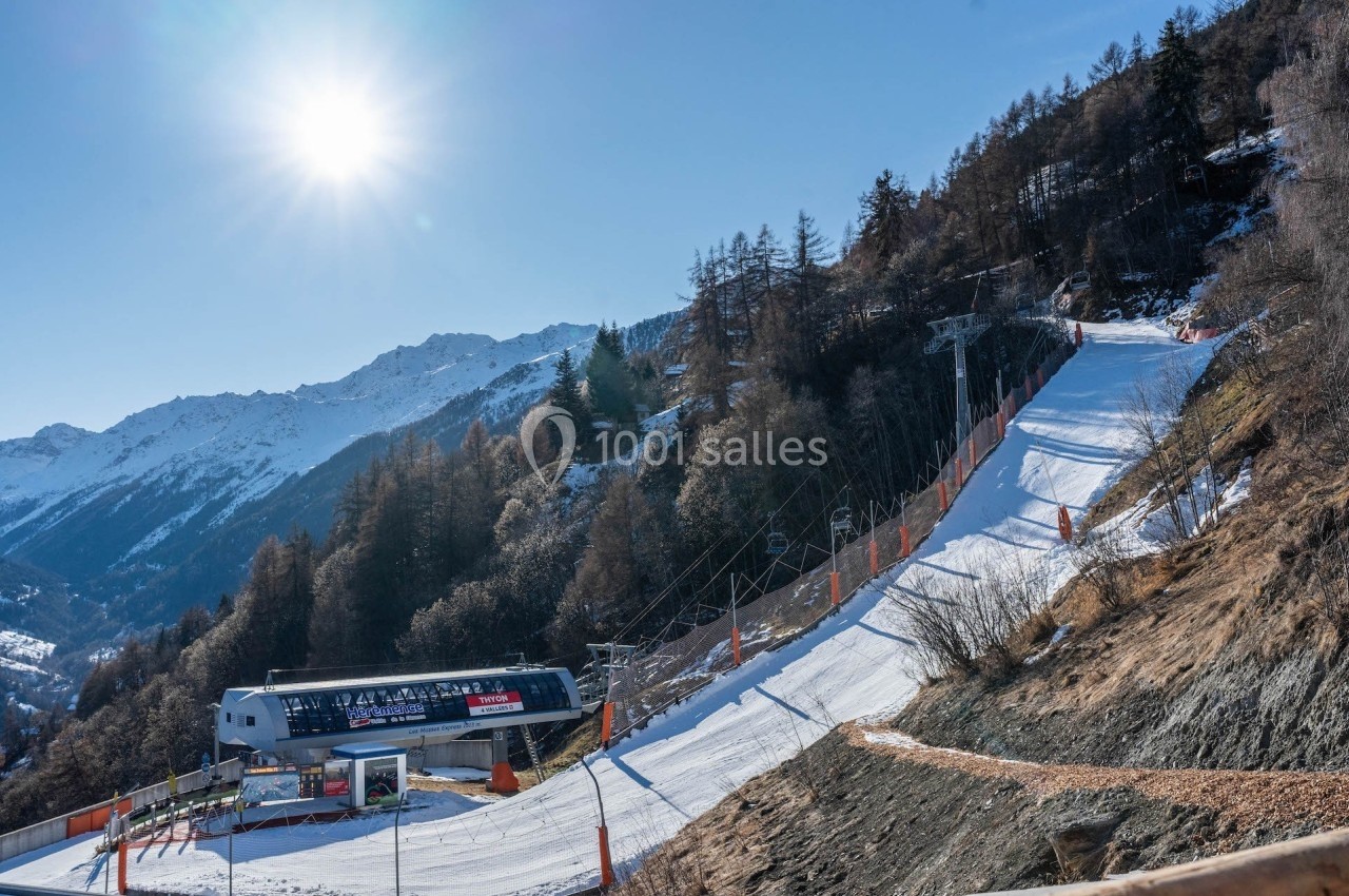 Piste de ski ensoleillée bordée de sapins avec un télésiège et des montagnes en arrière-plan.