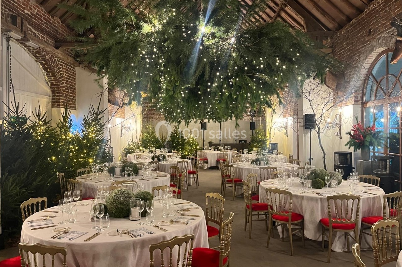 Salle de réception décorée pour un événement, avec tables rondes, guirlandes lumineuses et sapins ornant l'espace.