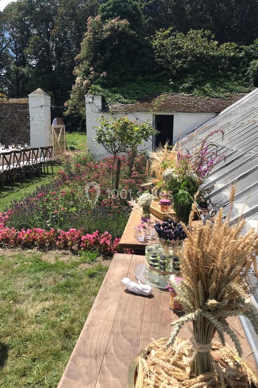 Table en bois décorée de fleurs et épis de blé, jardin fleuri et bâtiments anciens en arrière-plan.