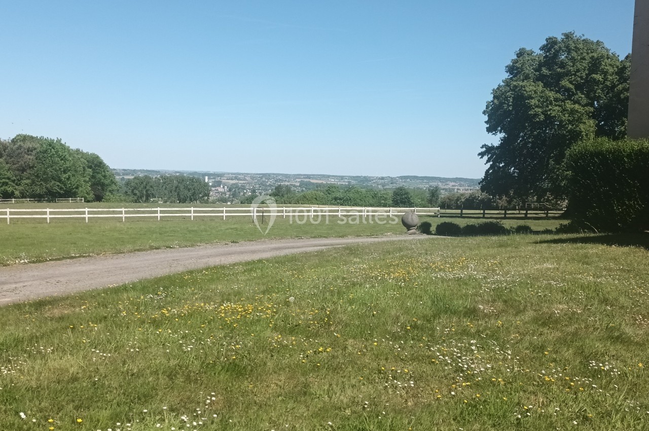 Vue d'un chemin bordé de pelouse parsemée de fleurs, avec des arbres, une clôture blanche et un paysage vallonné au loin.