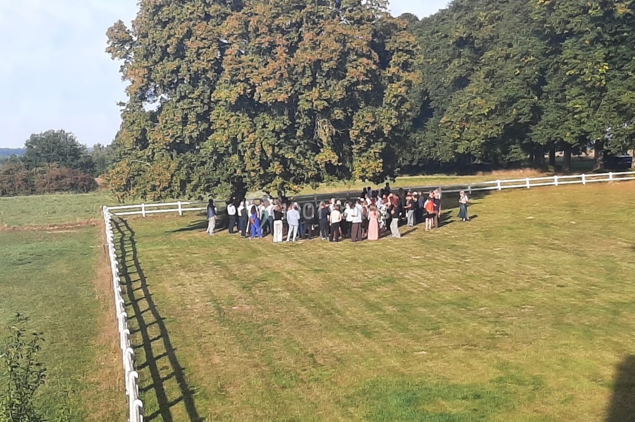 Groupe de personnes rassemblées sur une pelouse ensoleillée près d'une clôture blanche et d'arbres.