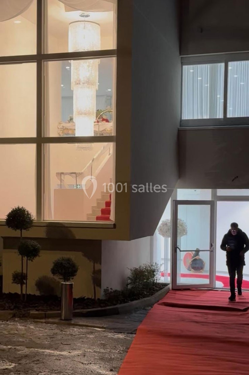 Entrée d'un bâtiment moderne avec escalier éclairé visible à travers une vitre et un homme marchant sur un tapis rouge.
