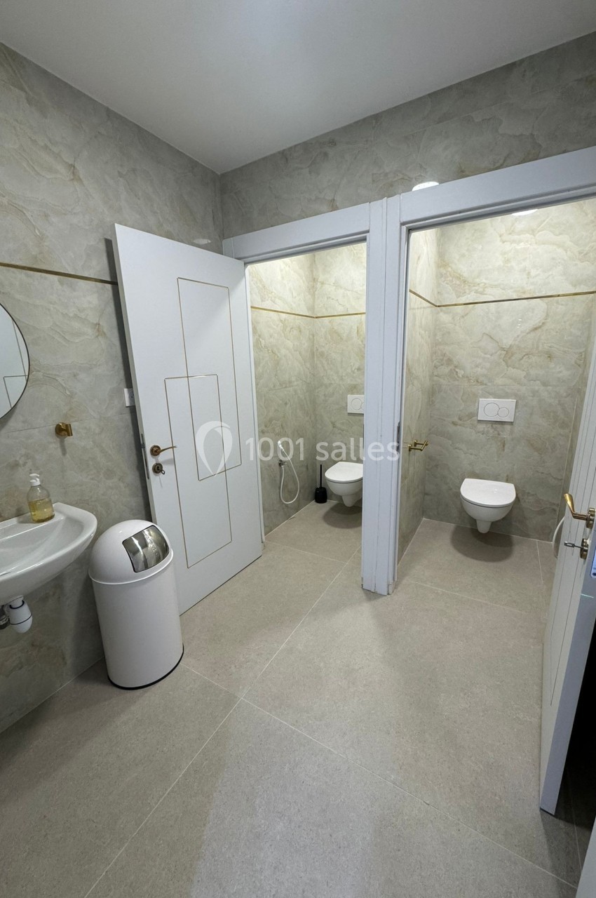 Salle de bain avec lavabo, miroir, poubelle et deux toilettes séparées par des cloisons.