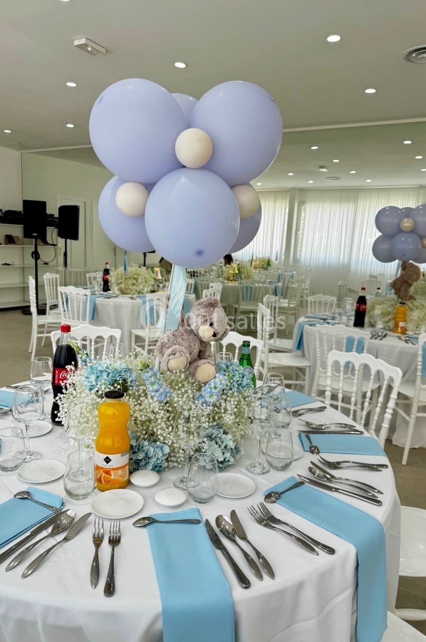 Table décorée pour un événement avec ballons violets, peluche, fleurs blanches et bleues, et couverts disposés.