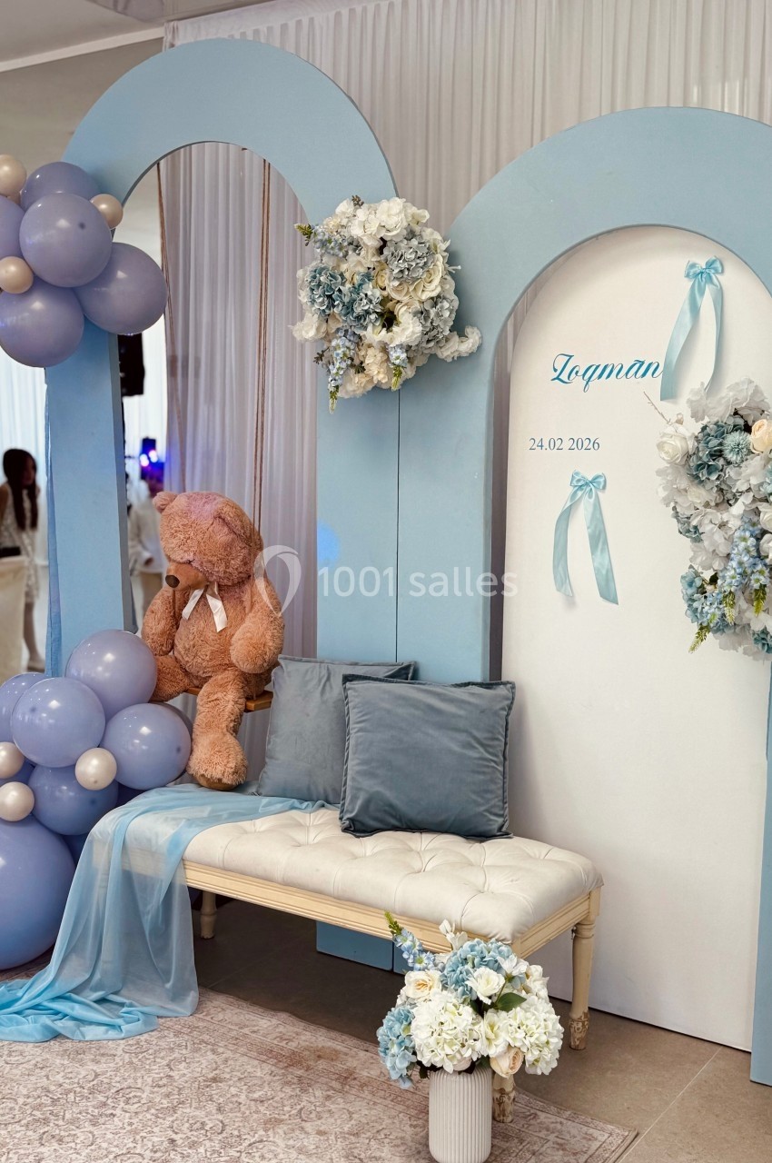 Décoration avec arches bleues, ballons, ours en peluche et fleurs, disposée autour d'un banc pour un événement.