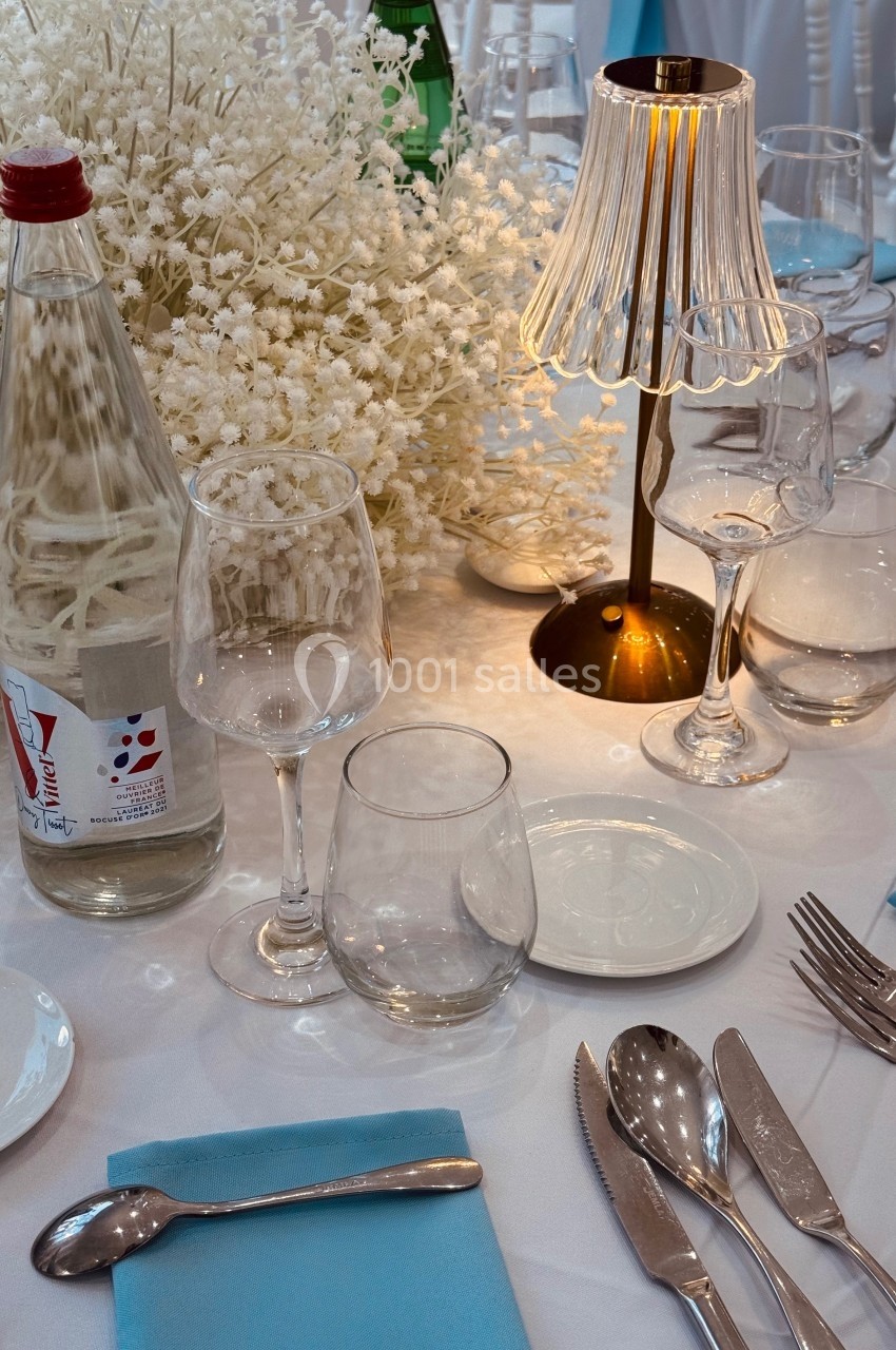 Table dressée avec nappe blanche, serviettes bleues, couverts, verres, bouteille d'eau et lampe près d'un bouquet blanc.