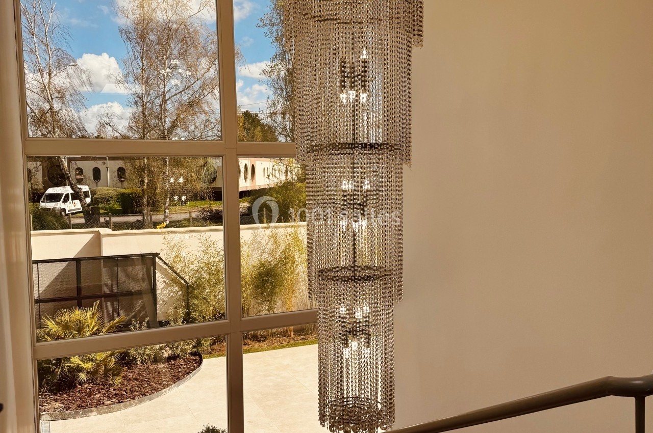 Lustre en cristal suspendu dans une cage d'escalier lumineuse avec vue sur un jardin et des bâtiments à travers une grande…
