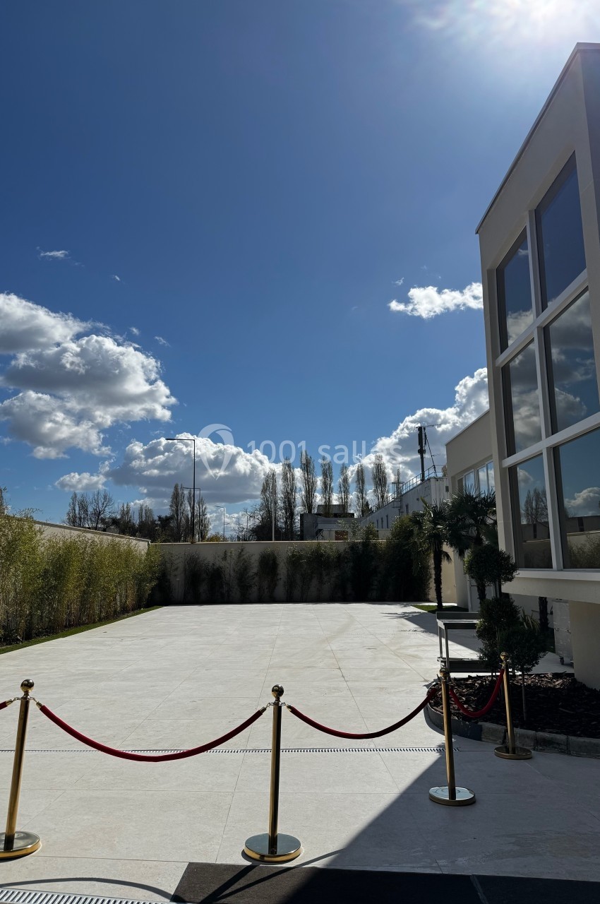 Cour extérieure ensoleillée avec sol en pierre claire, bordée de bambous et d'un bâtiment moderne aux grandes fenêtres.