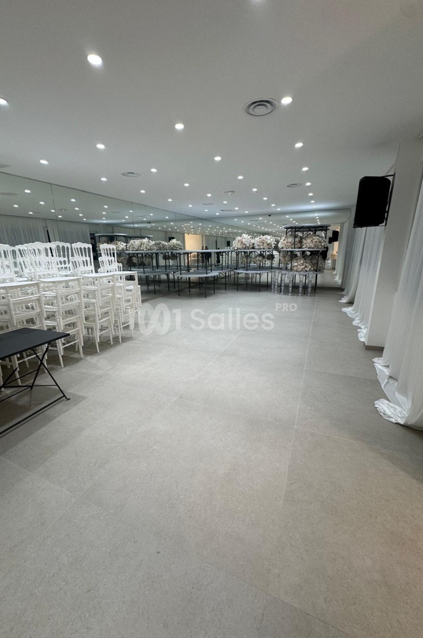 Salle de réception moderne avec sol en carrelage clair, chaises blanches alignées et tables décorées de fleurs.