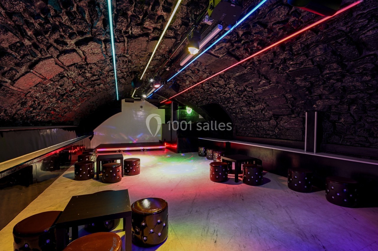 Salle voûtée en pierre avec éclairage néon coloré, tables basses et tabourets disposés sur un sol clair.