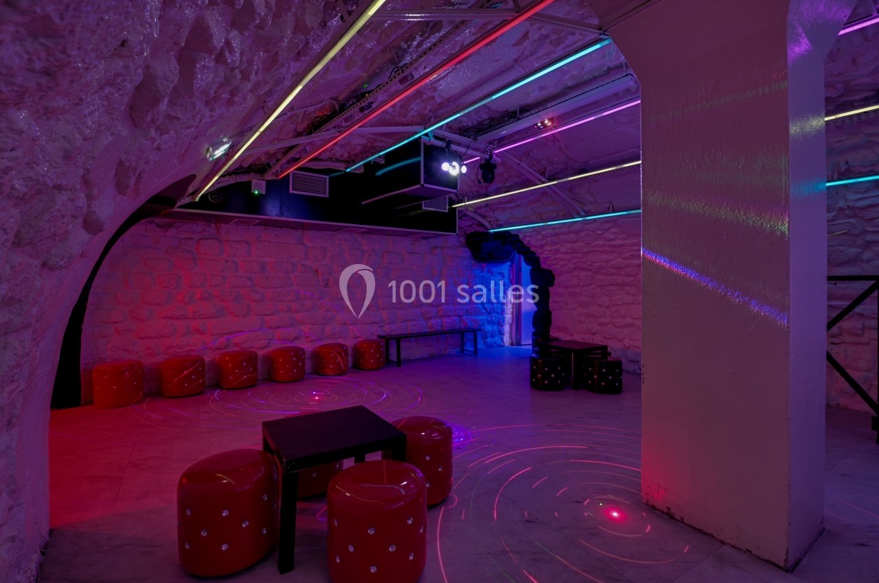 Salle voûtée en pierre avec éclairage néon coloré, tabourets rouges et tables noires disposés au sol.