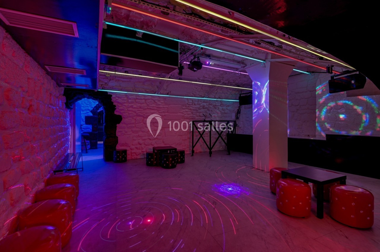Salle de club avec murs en pierre, éclairages colorés, tabourets rouges et piste de danse illuminée.