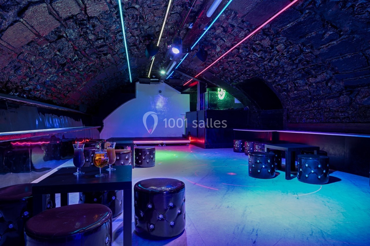 Salle voûtée avec éclairage néon coloré, tables basses, tabourets capitonnés et piste de danse vide.