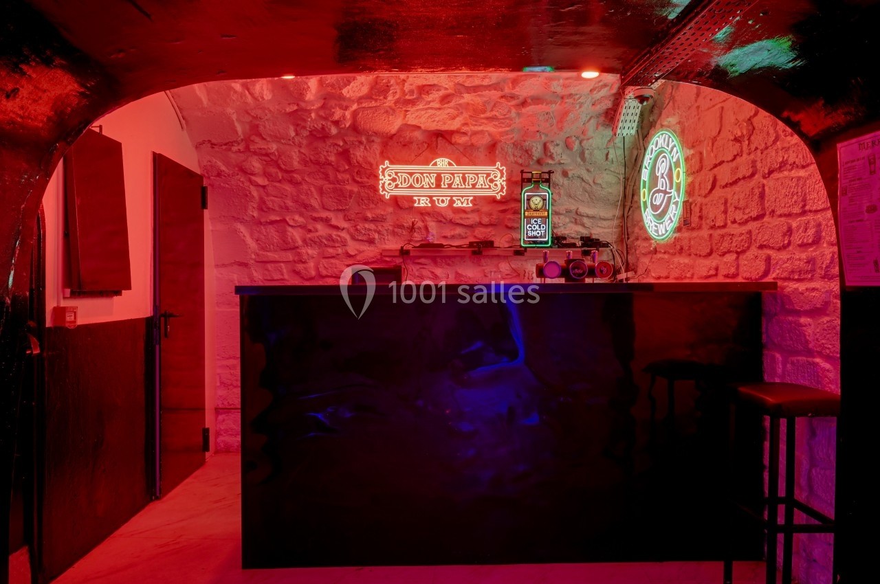 Bar avec comptoir noir, éclairé par des néons rouges et verts, dans une pièce aux murs en pierre apparente.