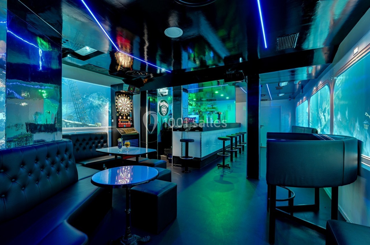 Intérieur d'un bar moderne avec éclairage néon, banquettes noires, tables basses et un jeu de fléchettes au mur.