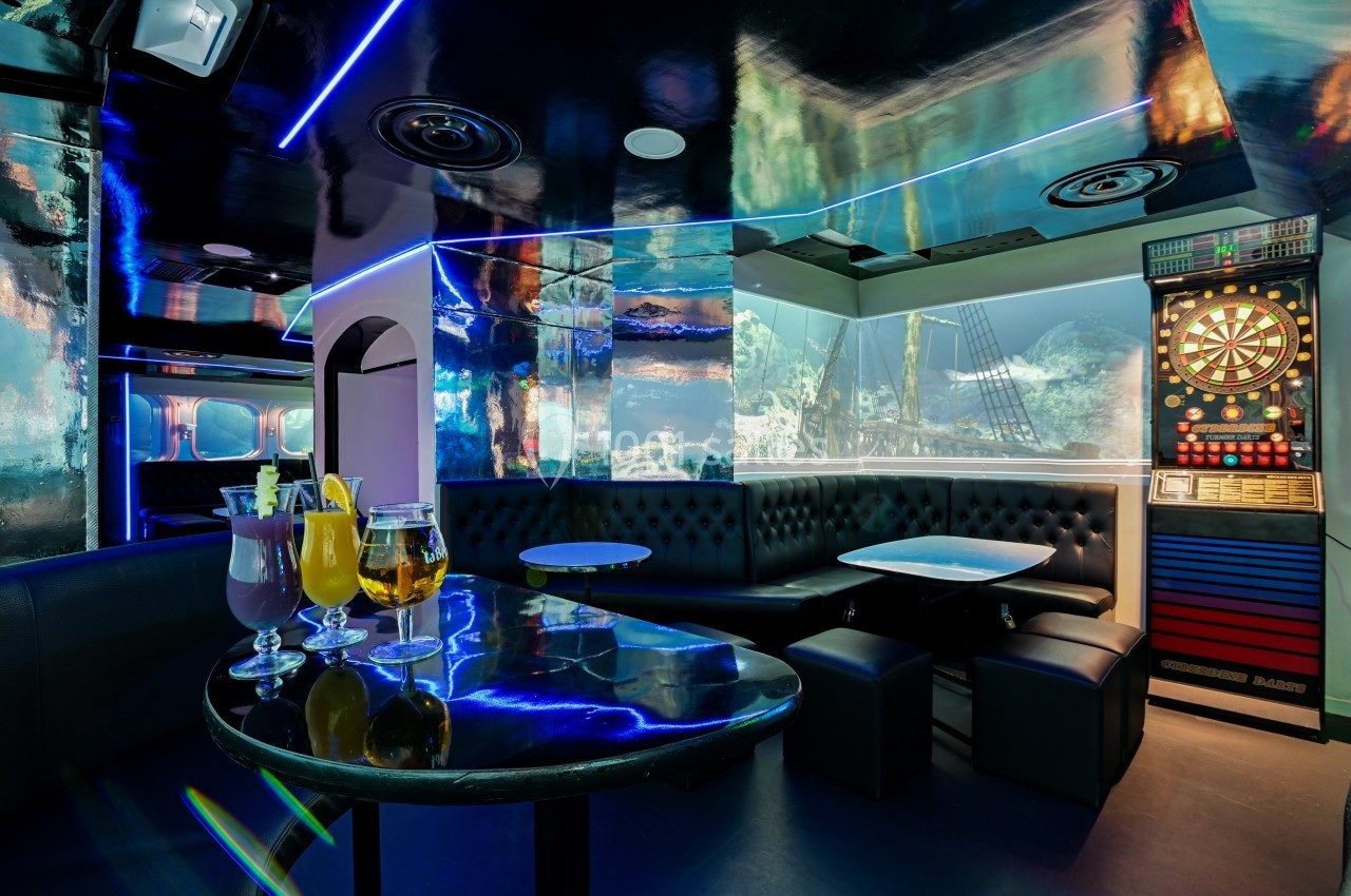 Salle de bar moderne avec banquettes noires, table basse, fléchettes électroniques et cocktails sur une table.