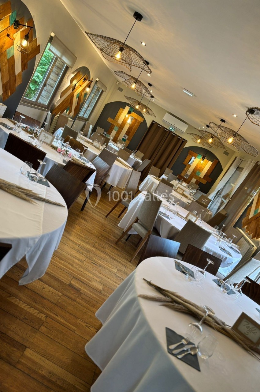 Salle de restaurant lumineuse avec tables rondes et rectangulaires dressées, décorée de bois et de luminaires suspendus.