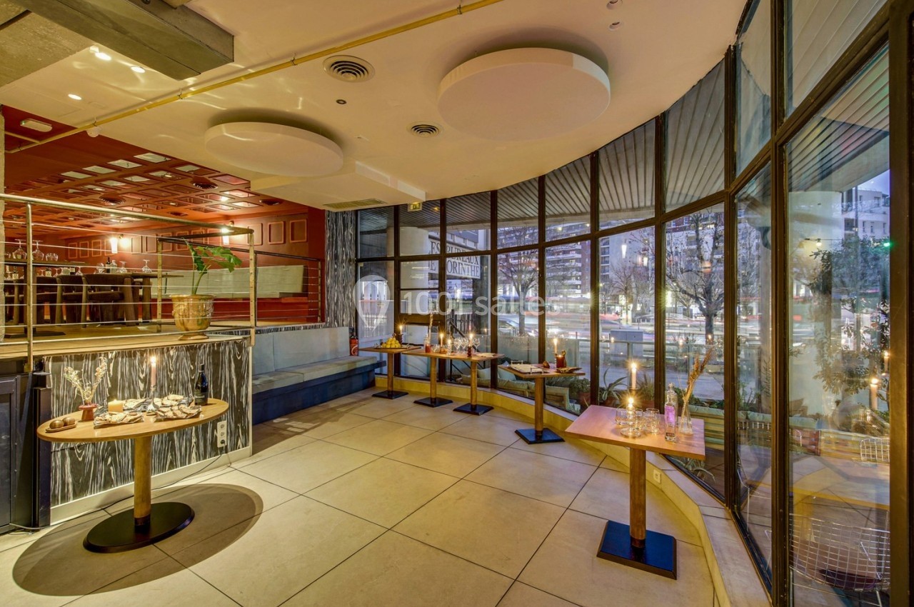 Salle de restaurant avec de grandes baies vitrées, tables dressées et vue sur un espace extérieur éclairé.