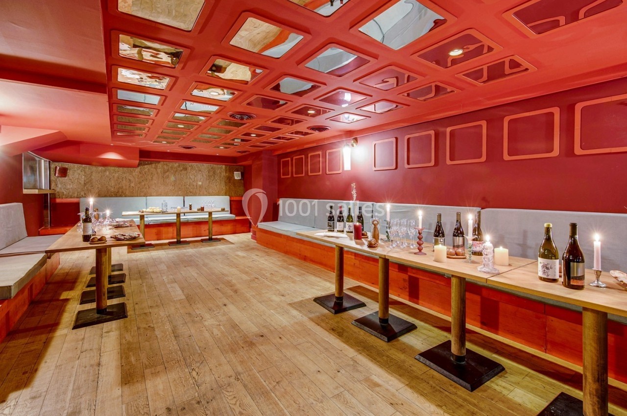 Salle avec tables en bois, bouteilles de vin, verres et bougies, éclairée par une lumière chaude et un plafond rouge.