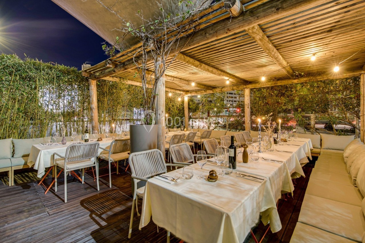 Terrasse d'un restaurant éclairée en soirée, avec tables dressées, chaises blanches et végétation en arrière-plan.