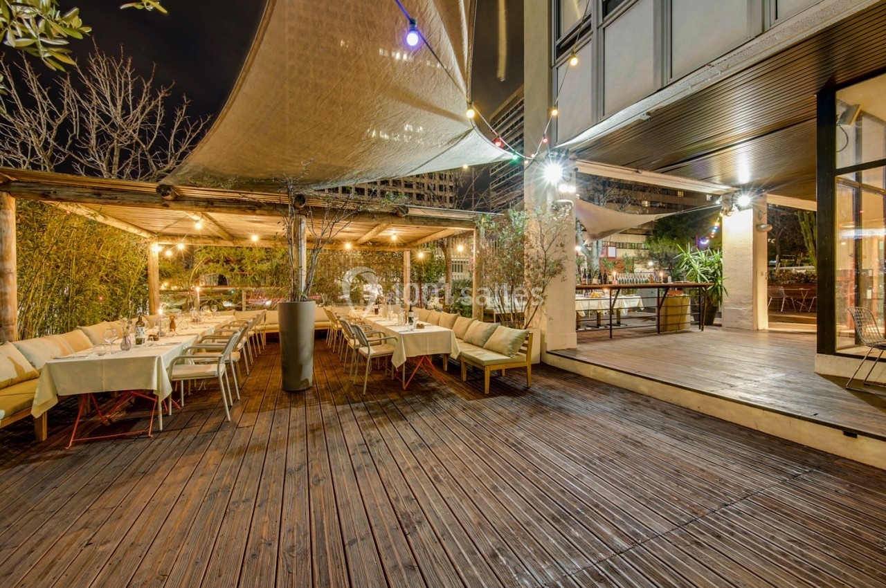 Terrasse en bois éclairée le soir, avec tables dressées sous une pergola et guirlandes lumineuses.