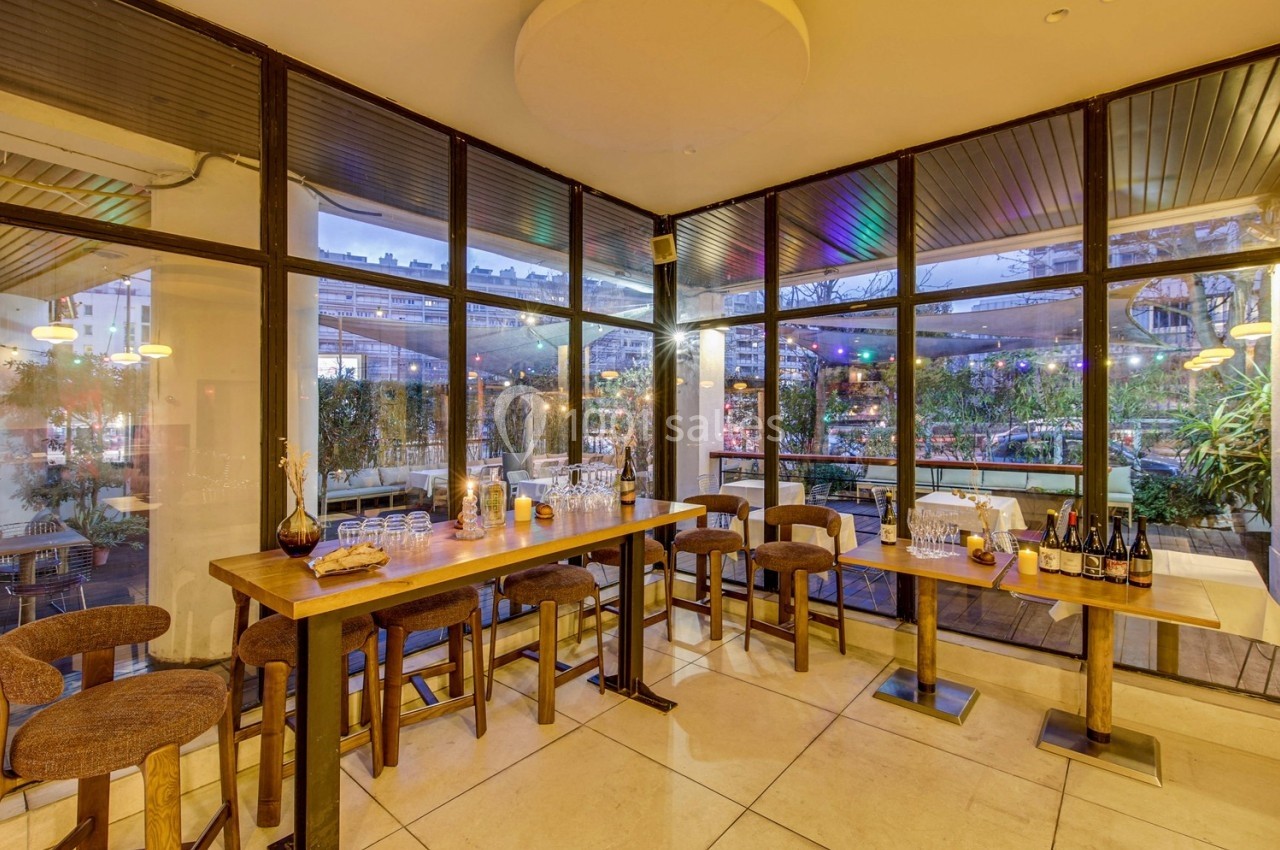 Salle de restaurant lumineuse avec de grandes baies vitrées, tables en bois, chaises hautes et vue sur une terrasse éclairée.