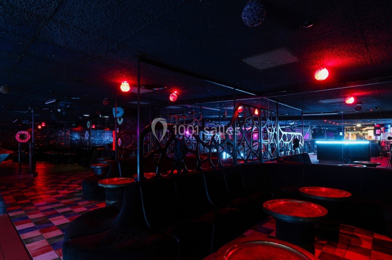 Salle de club avec éclairage rouge tamisé, banquettes noires, tables rondes et ambiance nocturne.