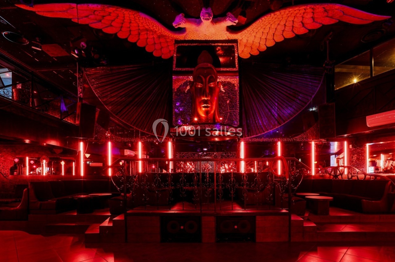 Salle de club avec éclairage rouge, décorations sombres, statue centrale et ailes illuminées en arrière-plan.