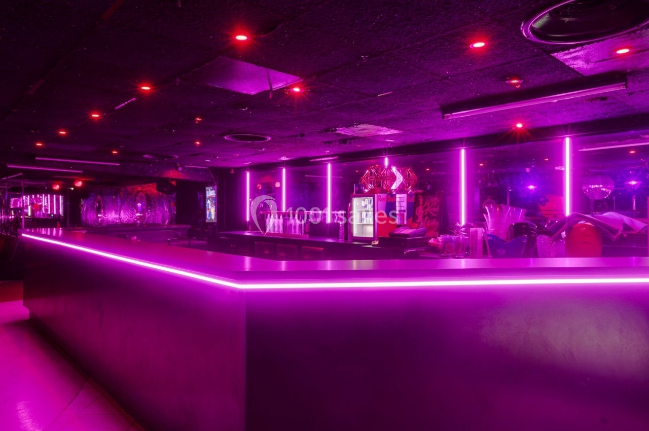 Bar avec éclairage néon rose, comptoir lumineux et rangées de bouteilles en arrière-plan dans une ambiance sombre.