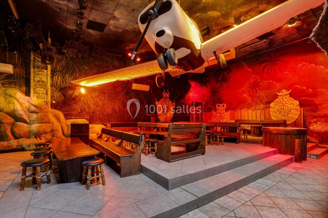 Salle à thème tropical avec un avion suspendu, des bancs en bois et une ambiance lumineuse rouge-orangée.