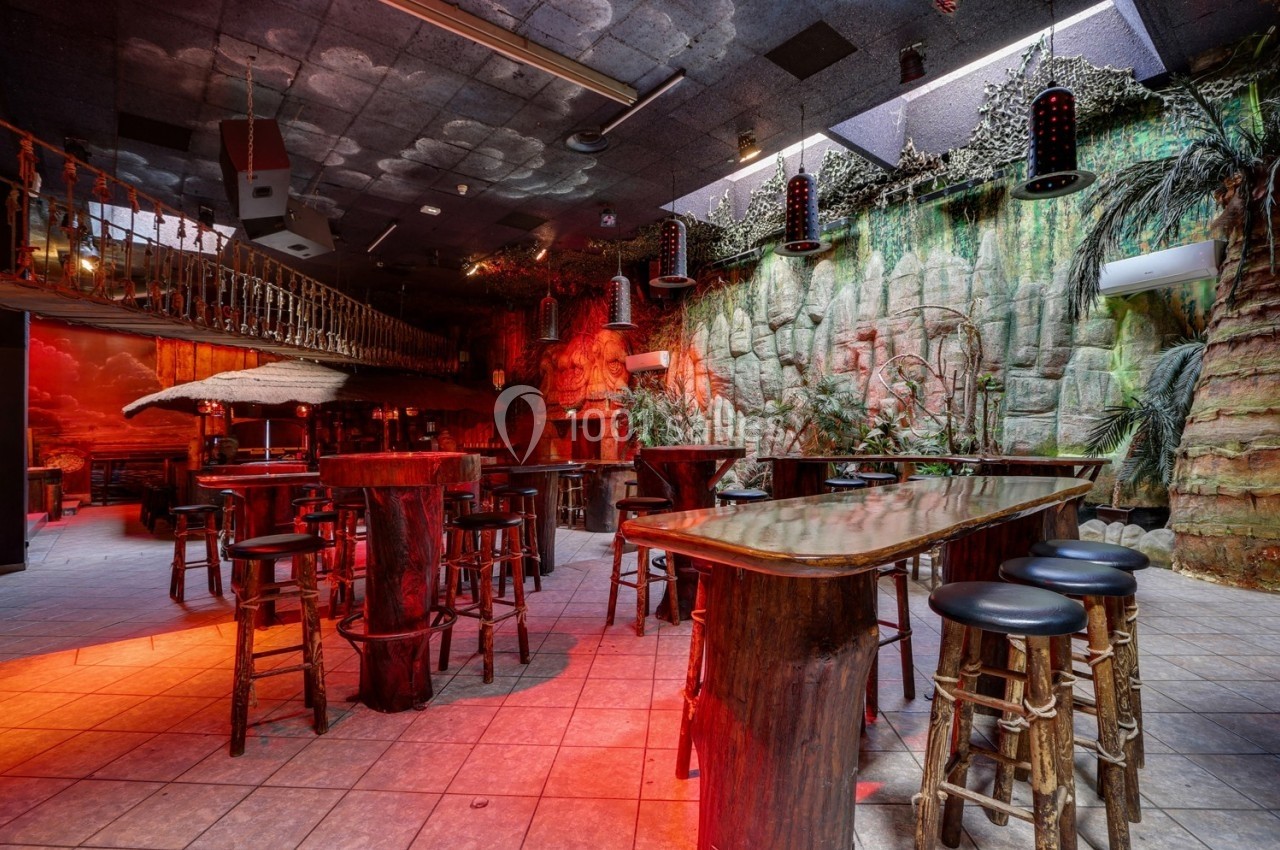 Salle de bar à thème tropical avec tables et tabourets en bois, éclairage rouge et décor végétal exotique.